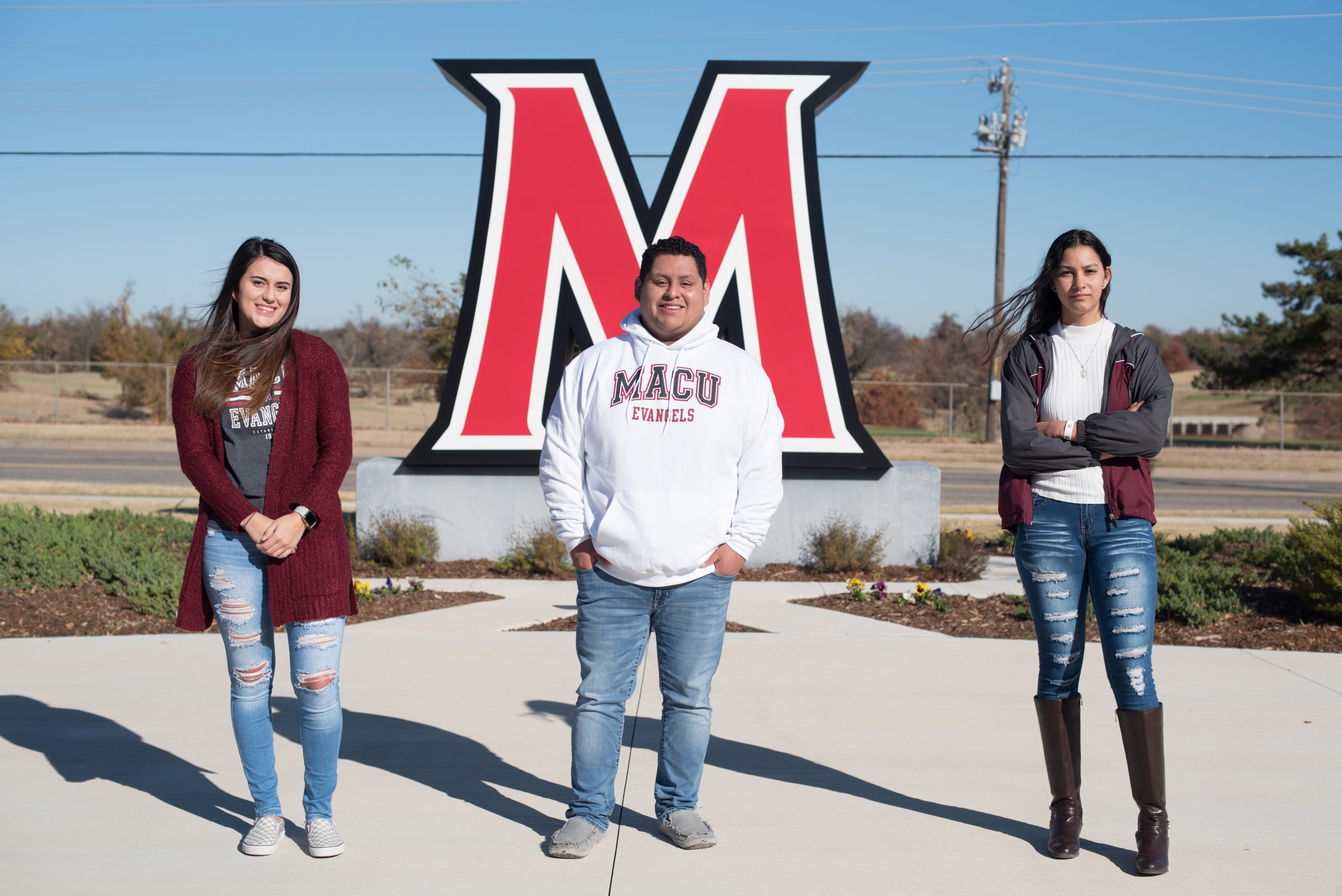 Two girls and one guy, wearing MACU apparel, in front of the "M" on campus.