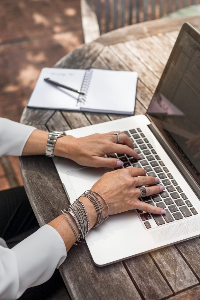 woman's hands typing on a laptop with notebook open beside her