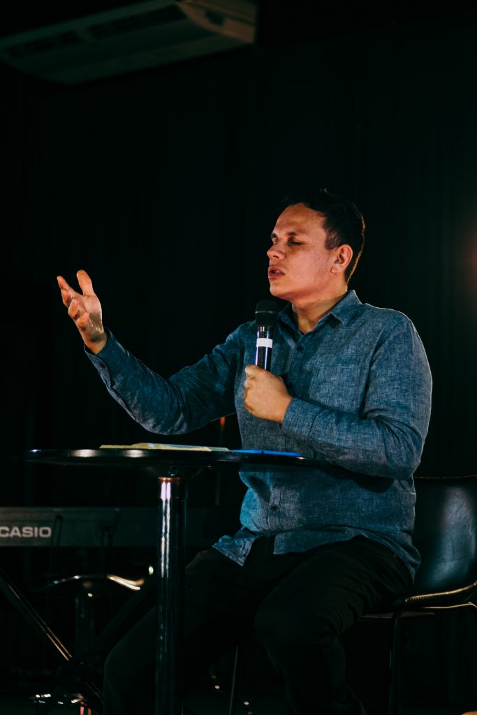 Young man sitting at small round table holding a microphone and praying.