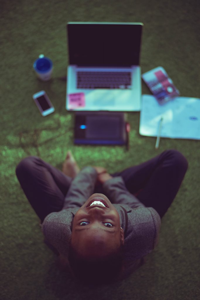 black woman sitting cross legged on floor in front of laptop, smiling up at camera