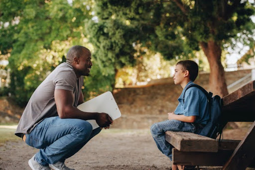 A school counselor squatting to talk to a student sitting on a bench outside.