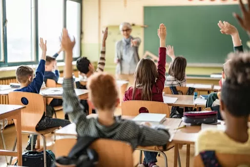 Students at their desks raise their hands in a classroom setting.
