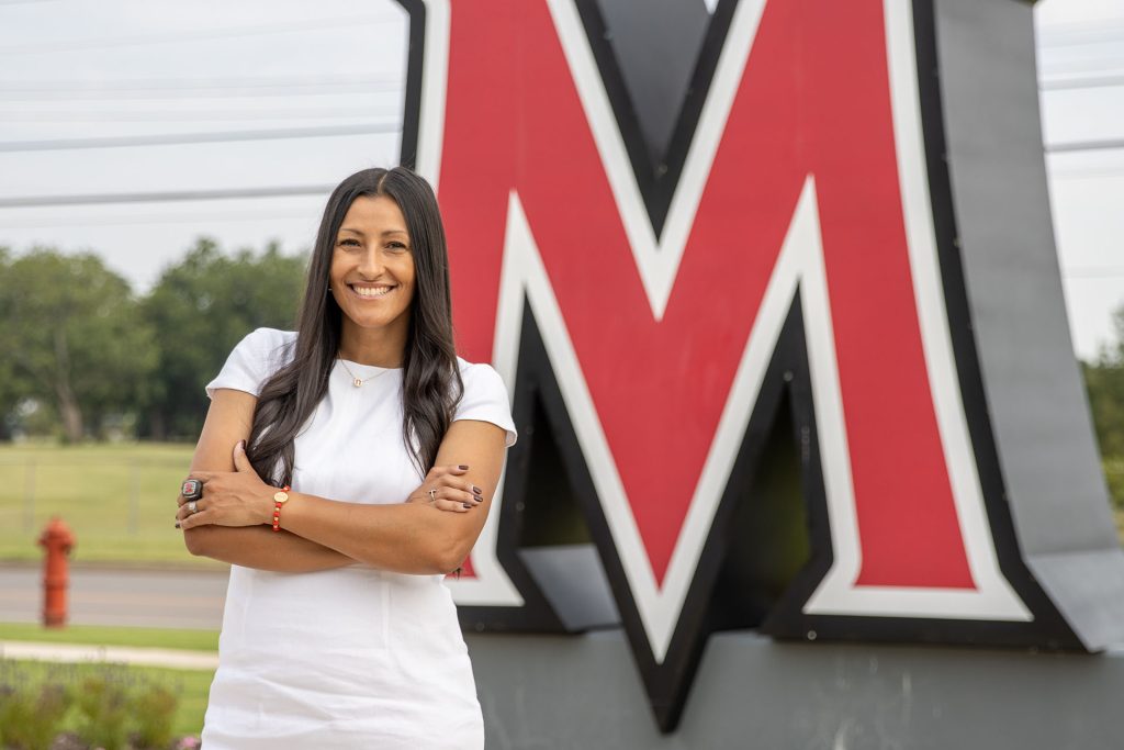 A woman in professional attire outside the MACU campus near a statue of the University logo.